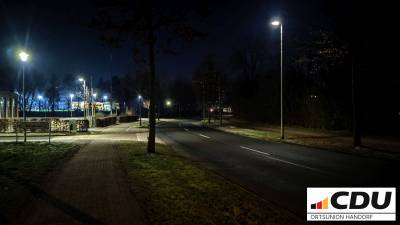 Hier sind die Zufahrt zum Bürgerbad und der Parkplatz des Feuerwehrgerätehauses aus Blickrichtung Dorbaum zu sehen. Die Beleuchtung ist besser als im südlicheren Bereich der Hobbeltstraße. Deutlich zu erkennen sind die Beleuchtung des Parkplatzes und des Sportplatzes im Hintergrund.